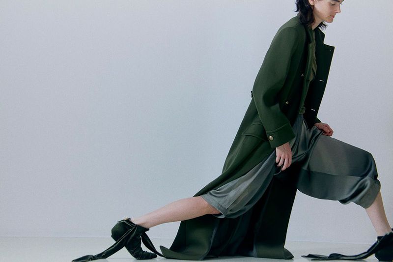 Fashion model wearing a long green coat and gray culottes with dark velvet ballet-style shoes posing in a brightly lit studio.