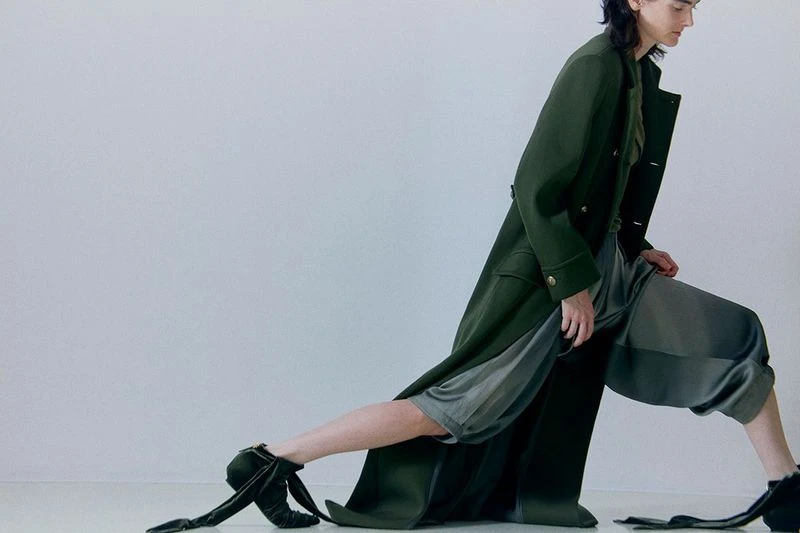 Fashion model wearing a long green coat and gray culottes with dark velvet ballet-style shoes posing in a brightly lit studio.