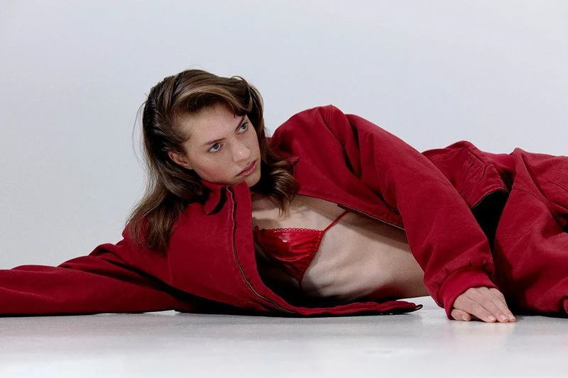 A woman in a red jacket and bra lies on her side in a studio setting with a clean, minimalist aesthetic, creating a striking visual.