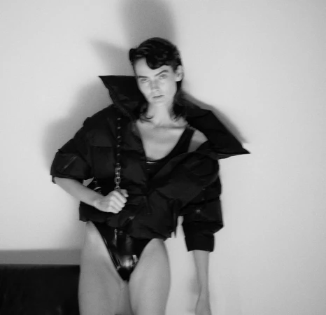 Monochromatic portrait of a model wearing a black puffer jacket and bodysuit against a plain wall in a photo studio setting.