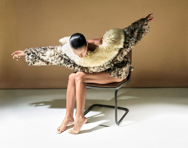 Woman in furry coat sitting on a chair in a studio with tan background. Barefoot, arms extended, she poses in the bright light.