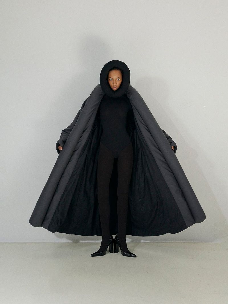 A woman in black bodysuit and black heeled boots wears an open dark grey puffer coat with a hood, posing in a white studio setting.