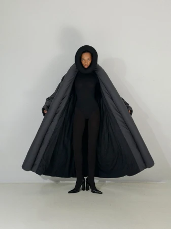 A woman in black bodysuit and black heeled boots wears an open dark grey puffer coat with a hood, posing in a white studio setting.