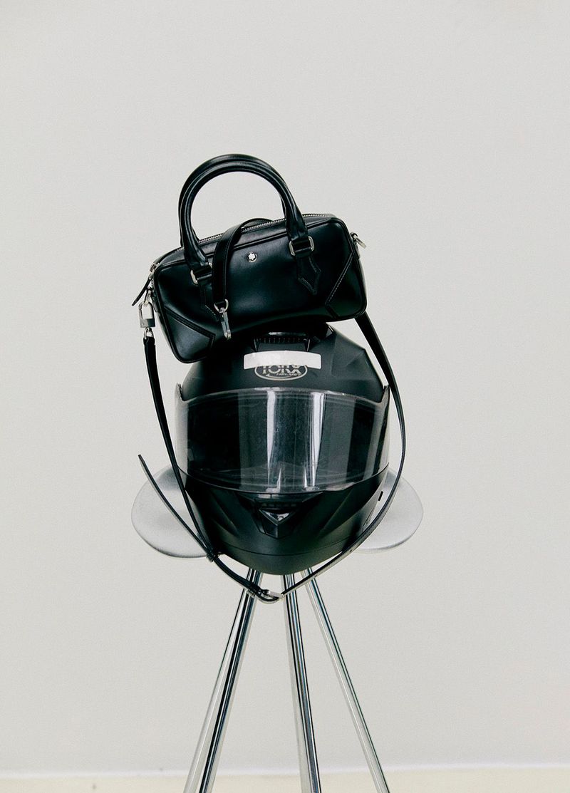Fashion still life: a black leather purse sits atop a sleek black motorcycle helmet on a chrome tripod stool against a white backdrop.