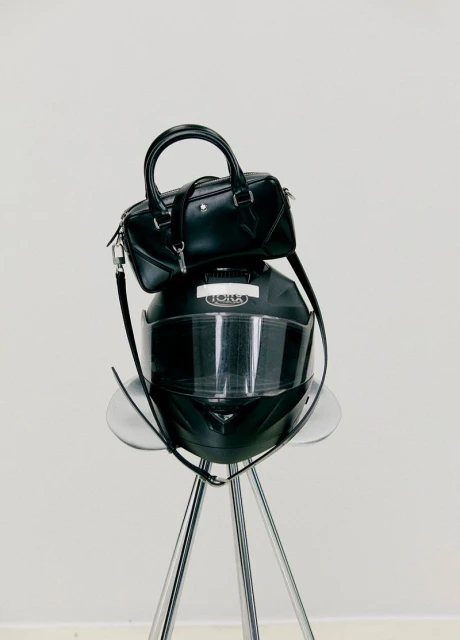 Fashion still life: a black leather purse sits atop a sleek black motorcycle helmet on a chrome tripod stool against a white backdrop.