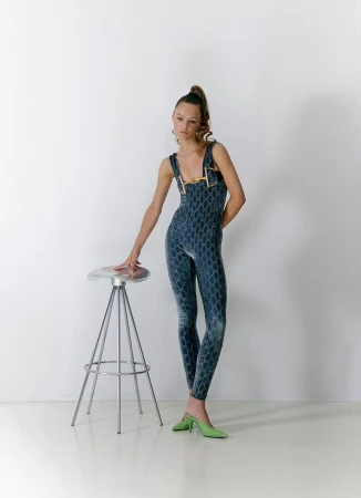 Adult female fashion model poses in a patterned, dark blue jumpsuit with pale green heeled shoes next to a silver stool in a studio setting.