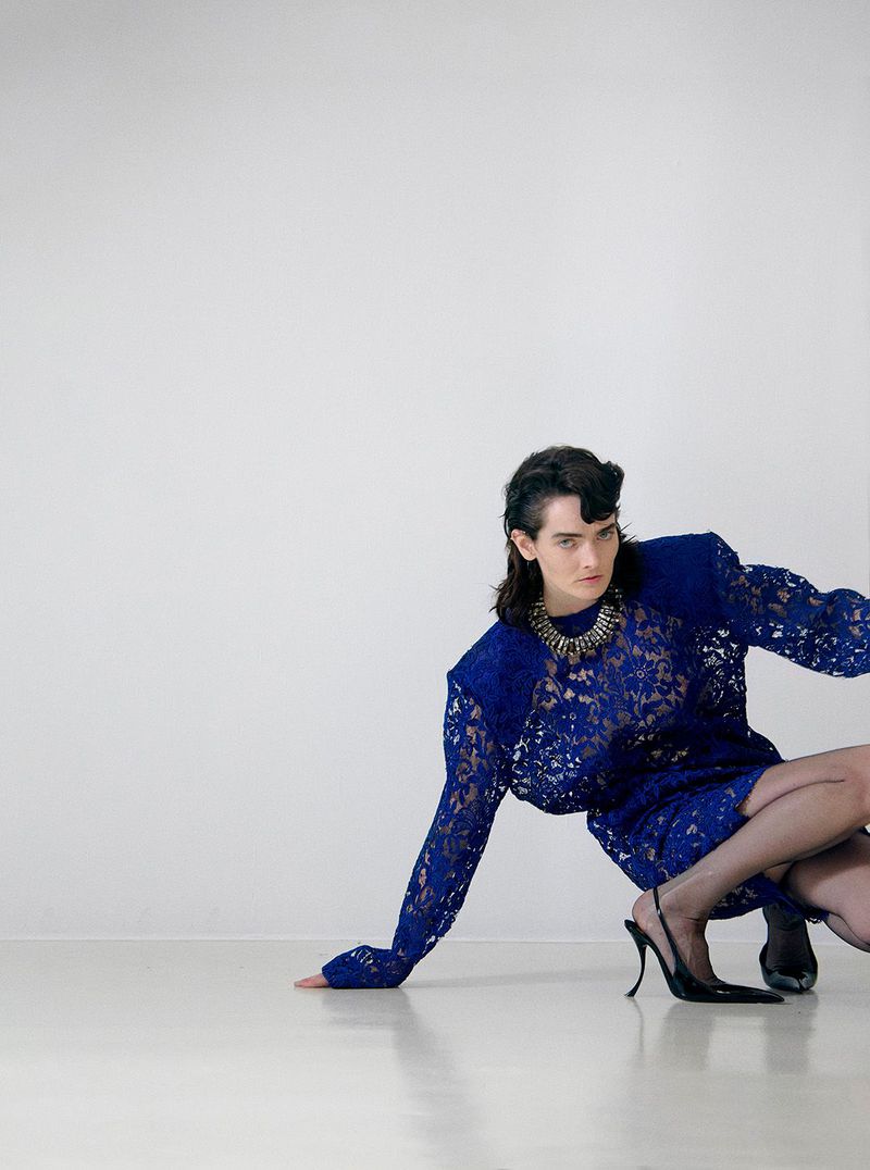 A person in a blue lace dress and black heels poses in front of a white wall, with the focus on the dress and the person's confident posture.