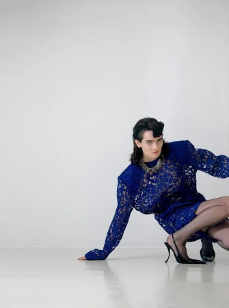 A person in a blue lace dress and black heels poses in front of a white wall, with the focus on the dress and the person's confident posture.