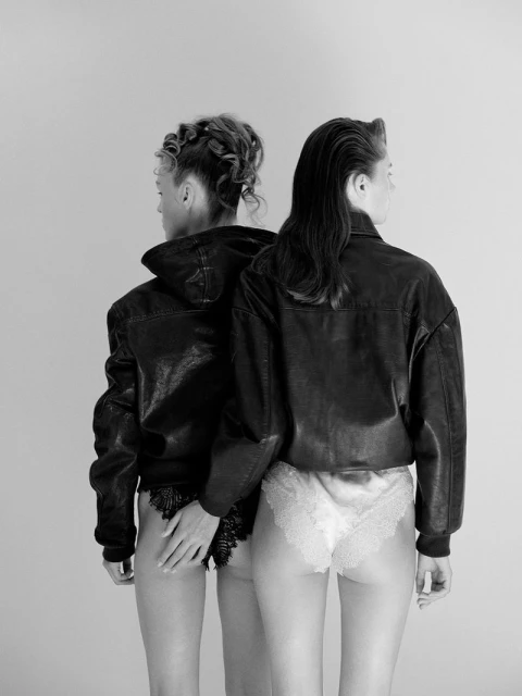 Back view of two women in underwear and leather jackets, one with hand on other's hip, in a black and white studio shot.