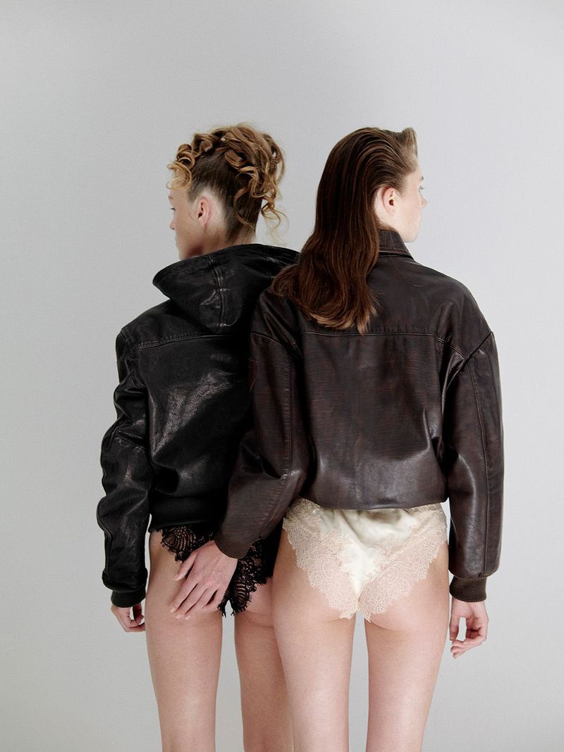 Two women pose back to back in leather jackets and lace lingerie against a neutral backdrop in a fashion forward studio setting.