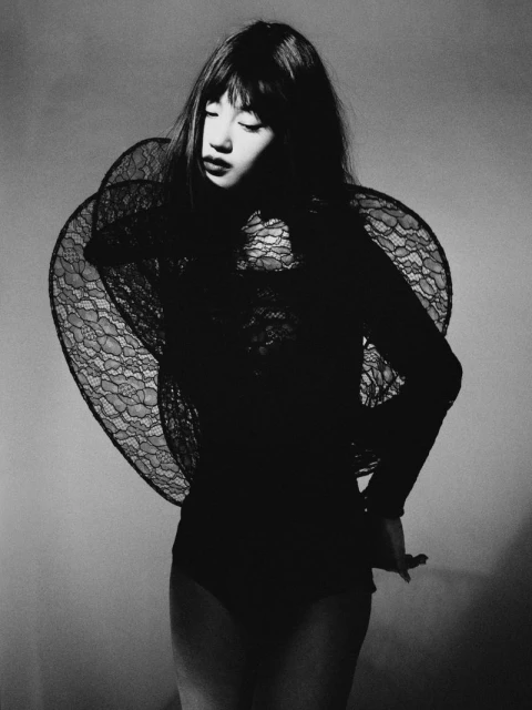 Monochrome studio portrait of a woman with bangs wearing a black bodysuit with lace shoulder detail posing in front of a grey background.