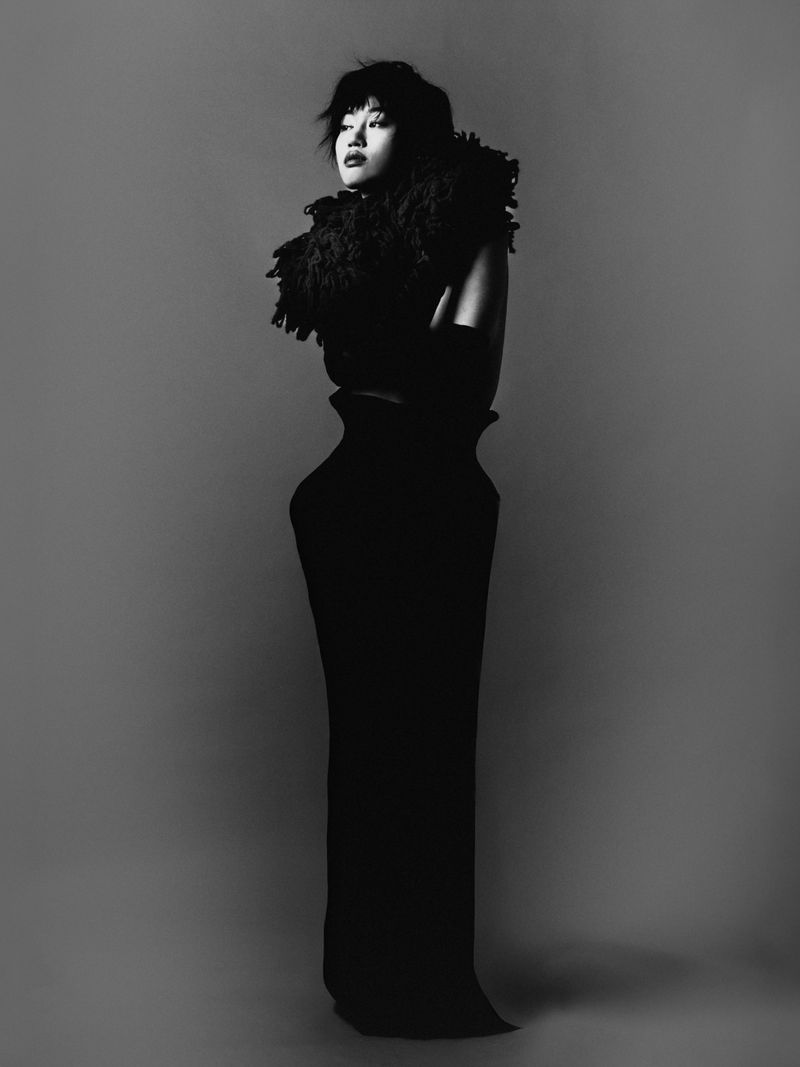 Monochrome studio portrait of an Asian woman in a long black dress with feather boa, standing with arms crossed against a grey background.