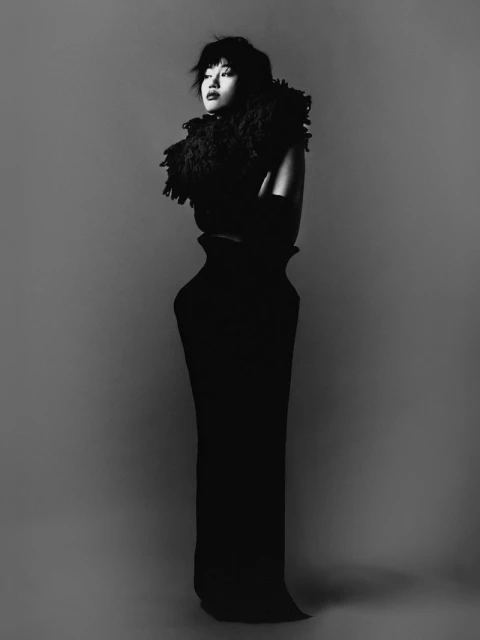 Monochrome studio portrait of an Asian woman in a long black dress with feather boa, standing with arms crossed against a grey background.