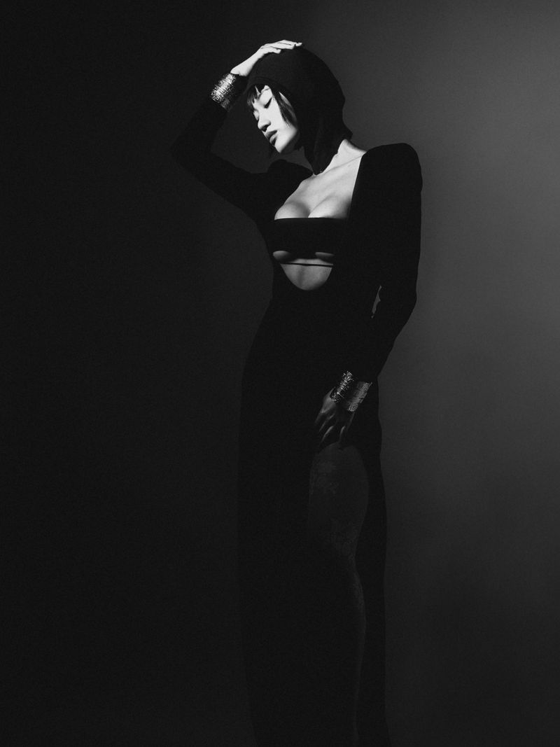 Monochrome portrait of a woman in a dark dress with cutouts, posing in a studio with a dark background, wearing metallic bracelets.