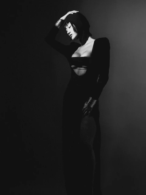 Monochrome portrait of a woman in a dark dress with cutouts, posing in a studio with a dark background, wearing metallic bracelets.