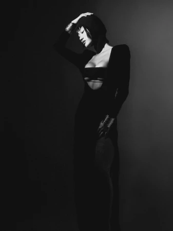 Monochrome portrait of a woman in a dark dress with cutouts, posing in a studio with a dark background, wearing metallic bracelets.