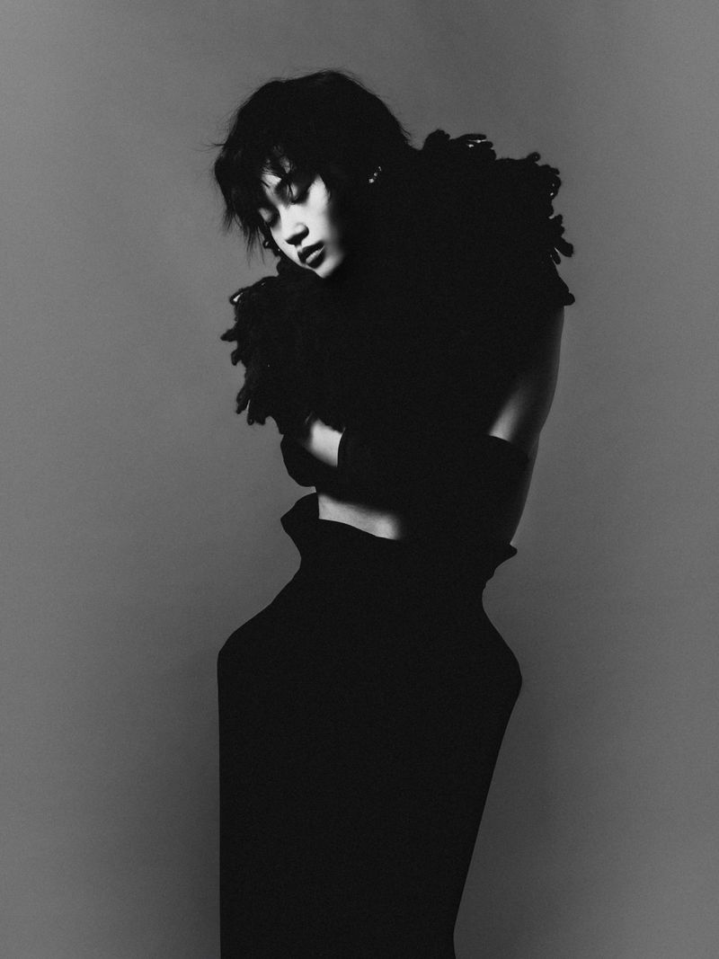 Monochrome portrait of a woman in an elegant black outfit with striking textures, against a muted grey backdrop in a studio setting.