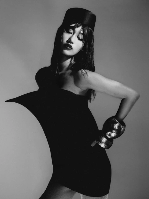 Black and white studio fashion portrait of an adult female model wearing a black sculptural dress and hat, with silver cuff bracelets.