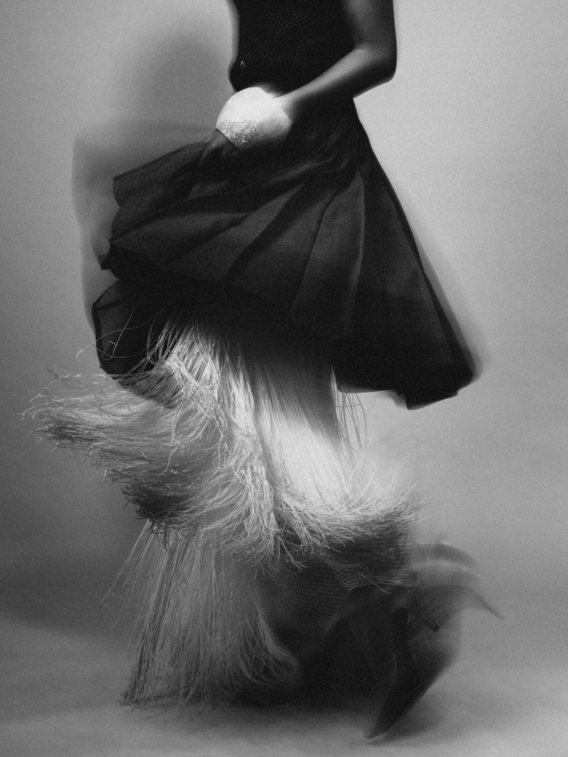 Monochrome image of a Black model in motion wearing a black dress with a flowing, fringed white skirt, against a soft grey background.