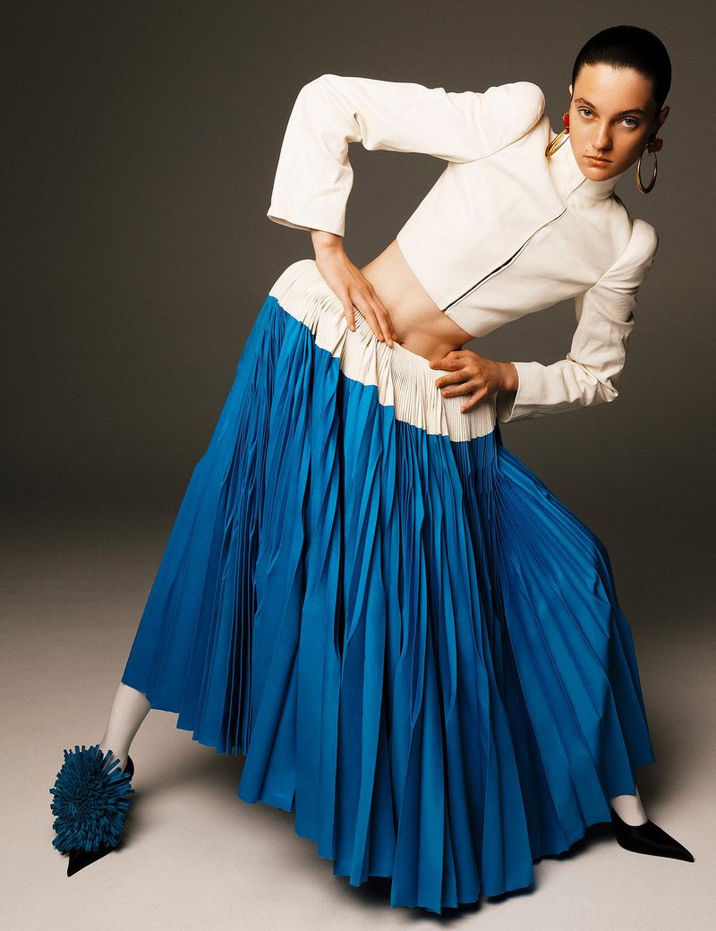 Fashion shot of a woman in a cropped white top and blue pleated skirt, posing with hands on hips against a grey studio backdrop.