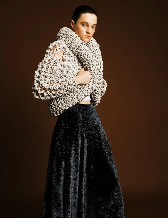Woman with short hair wearing a cream knitted jacket and a shiny black skirt posing in front of a brown background in a studio shot.