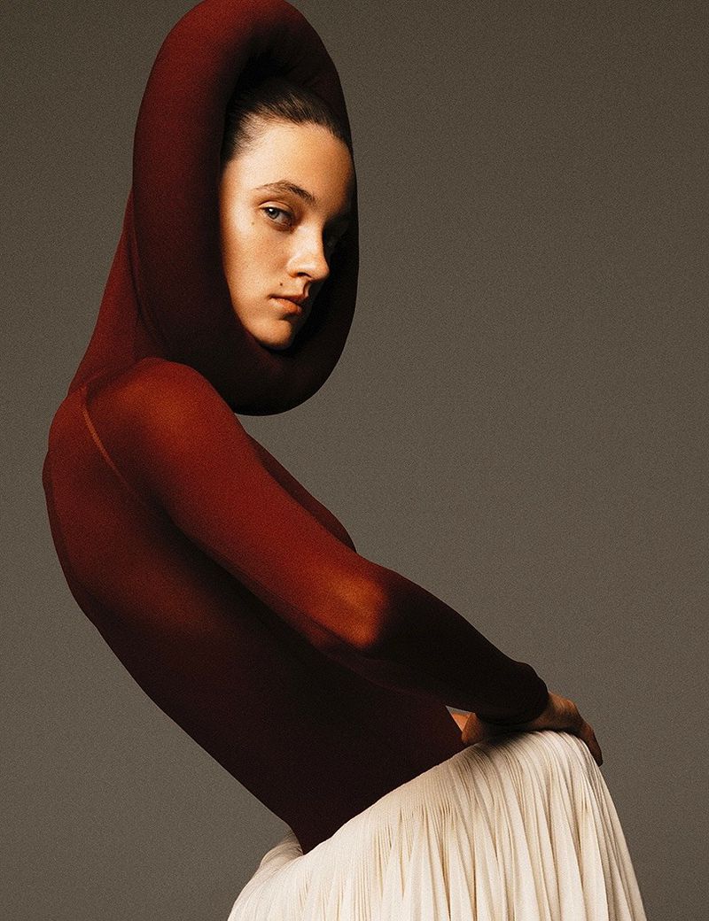 Woman in a sculptural hooded garment and cream-colored skirt, facing forward, in a studio setting with neutral background and diffused lighting.