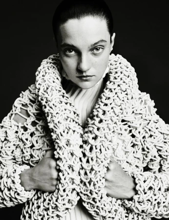 Monochrome portrait of an adult female with short dark hair wearing a white knit jacket. Direct eye contact on a dark background.