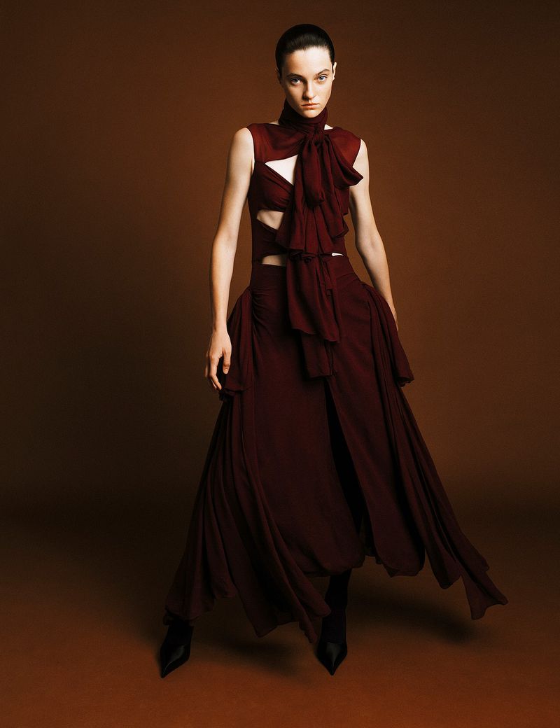 Woman in a dark red flowing gown with ruffles poses against a brown background, with dark hair slicked back in a fashion shot.