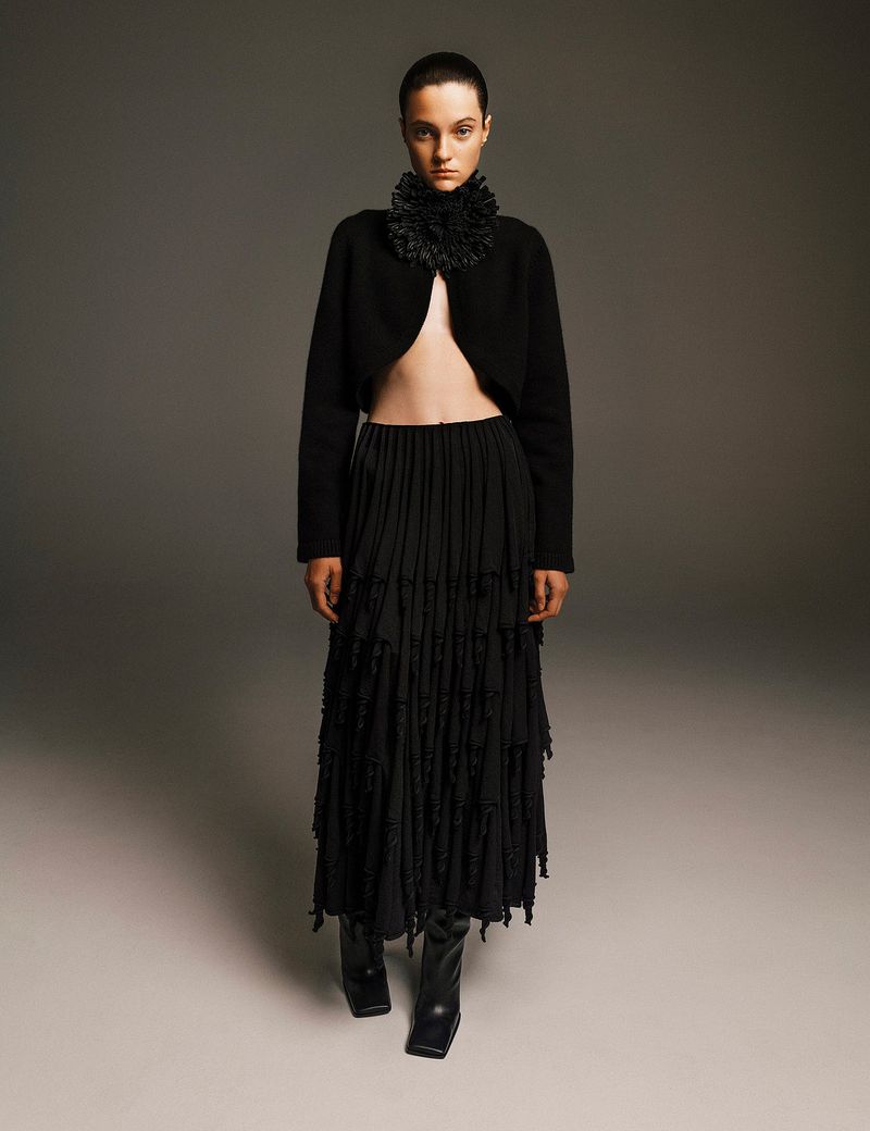 Elegant woman in black bolero jacket and tiered maxi skirt poses in a studio setting with neutral background and professional lighting.