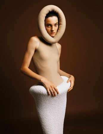 A woman poses in avant-garde fashion with a beige bodysuit, a white textured skirt, and a beige donut-shaped collar against a brown background.
