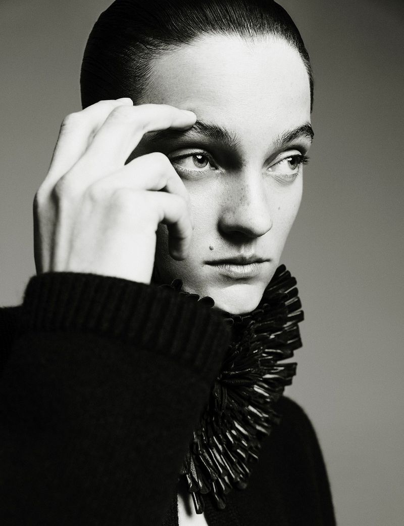 Monochrome portrait of a woman with hand on forehead, looking away, wearing a black top with a distinctive spiky collar.