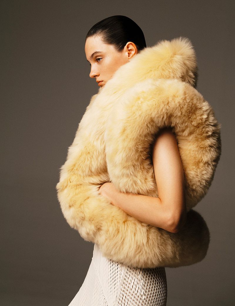 Fashion portrait of a stylish adult female wearing a large furry beige vest against a grey backdrop in a photo studio.