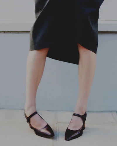 Cropped view of a person wearing a dark midi skirt and black pointed-toe Mary Jane low heels standing on a paved outdoor surface.
