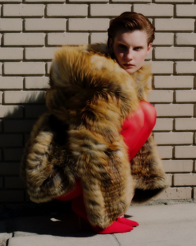 Model crouching against a brick wall wearing an oversized brown fur coat and vibrant red tights in direct sunlight.