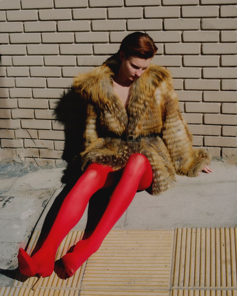 A woman with short red hair sits on a sidewalk in a fur coat and red tights against a brick wall, posing in bright afternoon sunlight.