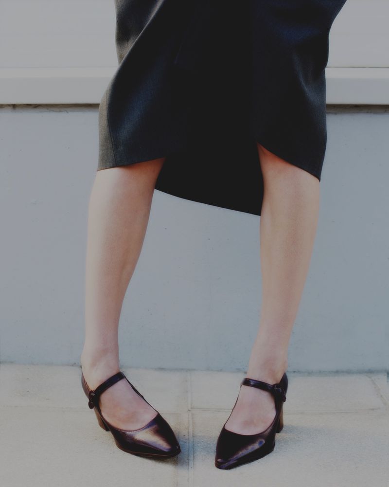 Cropped view of a person wearing a dark midi skirt and black pointed-toe Mary Jane low heels standing on a paved outdoor surface.