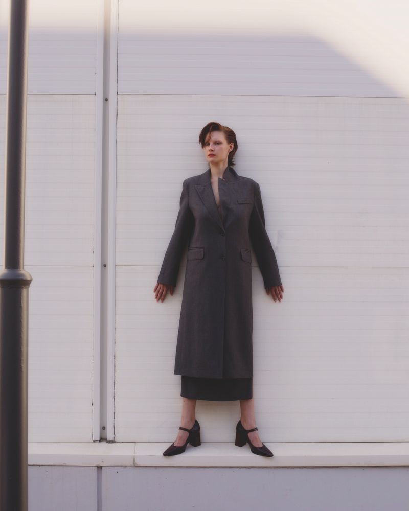 A woman in a gray coat and black shoes stands in front of a white wall with a black pole on the left, lit by sunlight.