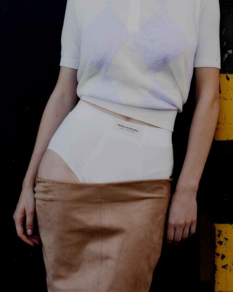 Mid-section portrait of a woman in white underwear, a tan suede skirt, and a white t-shirt posing against a black wall.