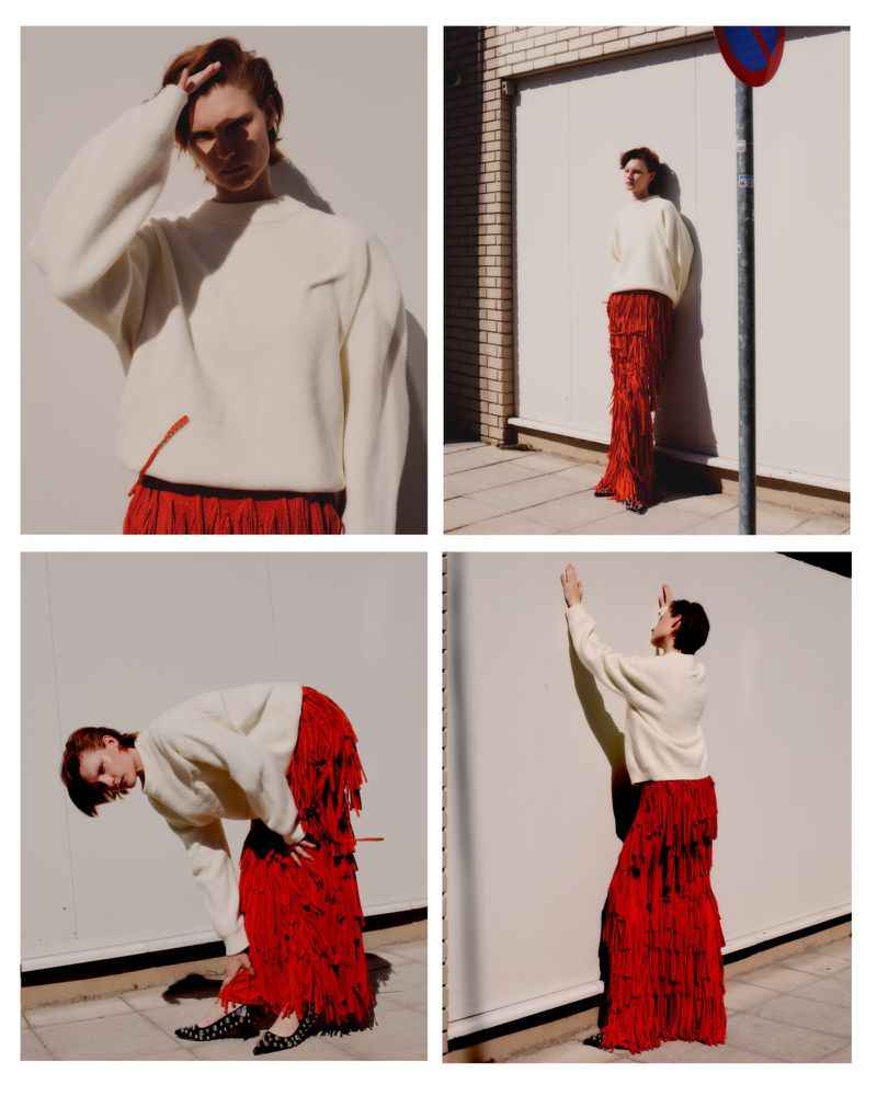 A woman posing in a cream sweater and red fringe skirt against a white wall in a fashion editorial style, showcasing a modern, chic aesthetic.