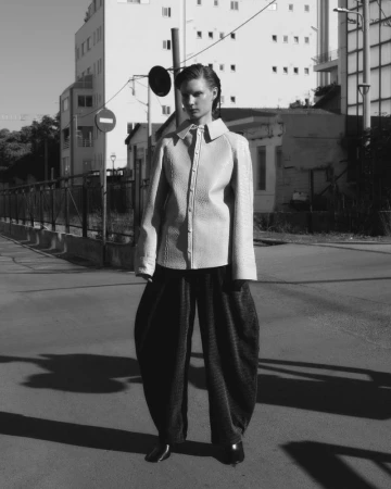 A woman in a patterned collared jacket and baggy trousers stands on a street, with buildings and road signs visible in the background.