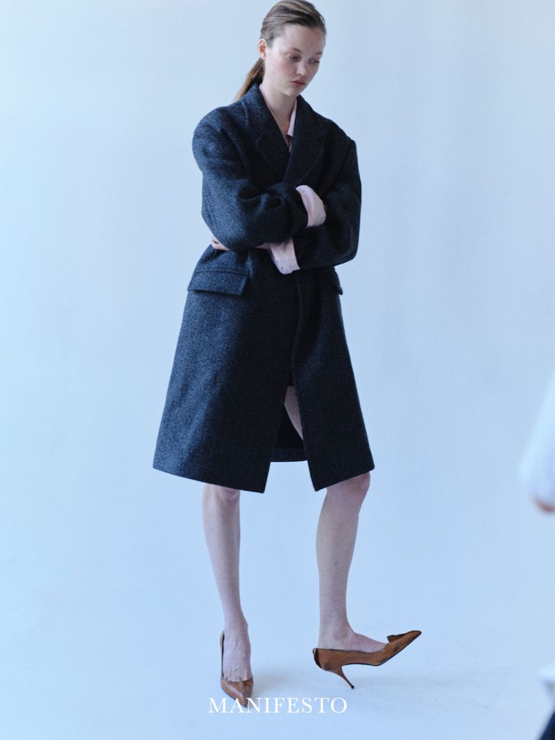 Woman poses in a navy coat and heels against a light background, highlighting fashion and studio photography with a modern, editorial aesthetic.