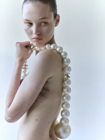 Woman poses nude with large Chanel pearl necklace in studio setting, artful fashion portrait with a focus on the jewelry.