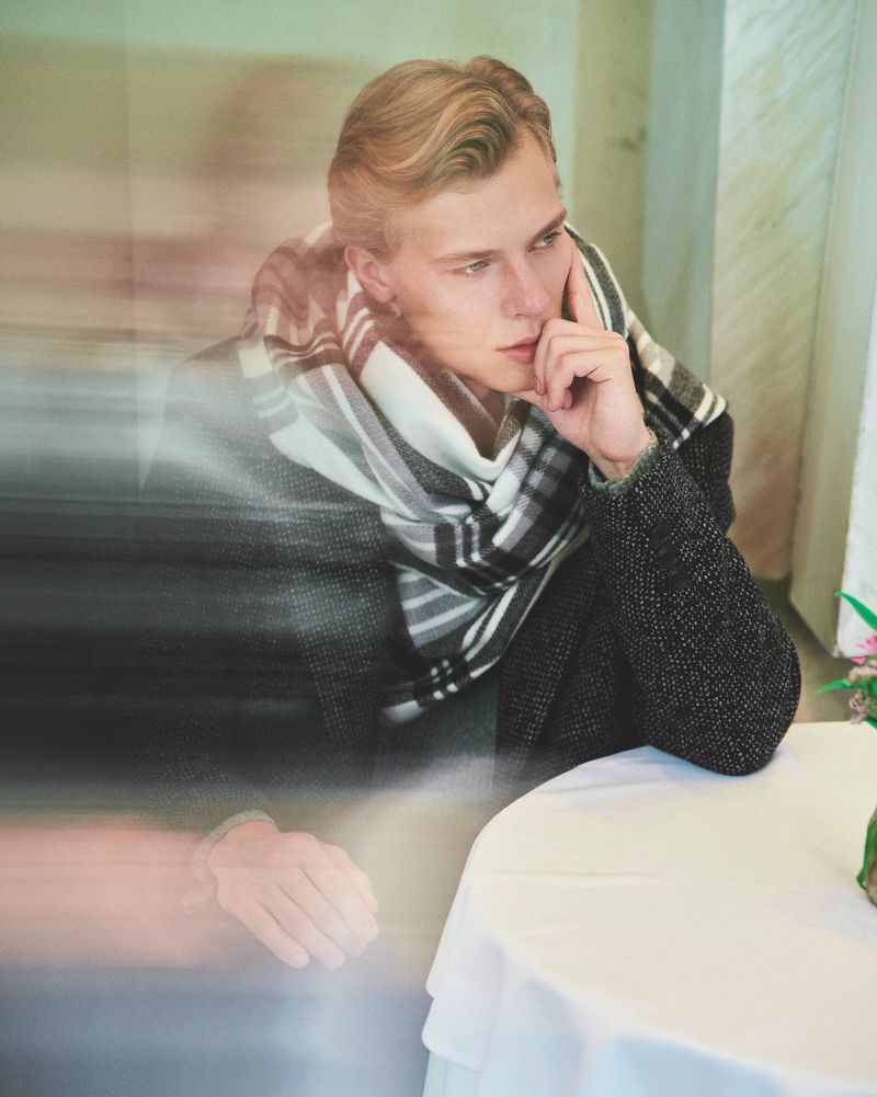 Portrait of a blond male in a coat and striped scarf seated at a table with a white tablecloth, with soft lighting and visual effects.
