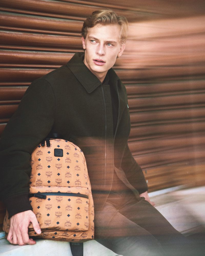 Handsome man with blond hair carrying an MCM backpack wearing a dark coat against a textured brown background, looking away.