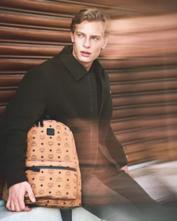 Handsome man with blond hair carrying an MCM backpack wearing a dark coat against a textured brown background, looking away.