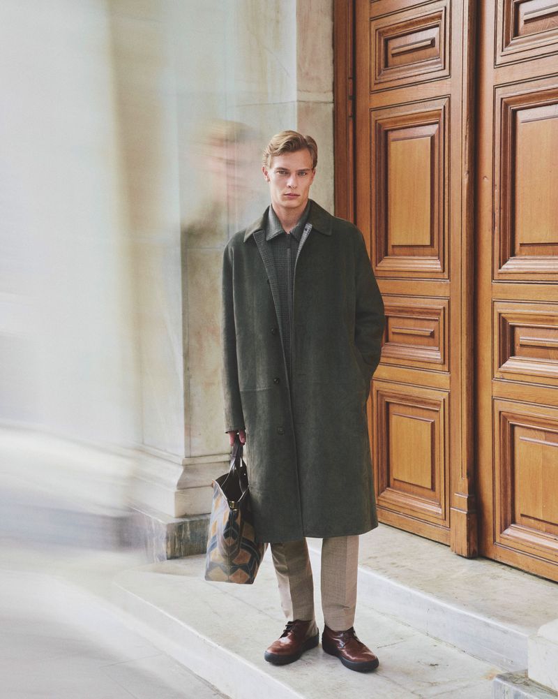 Adult male in a green overcoat, tan pants, and brown leather shoes stands next to a building with large wooden doors holding a patterned tote bag.