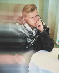 Blond man in a grey plaid scarf and dark coat seated at a café table, resting his chin on his hand with a motion blur overlay.