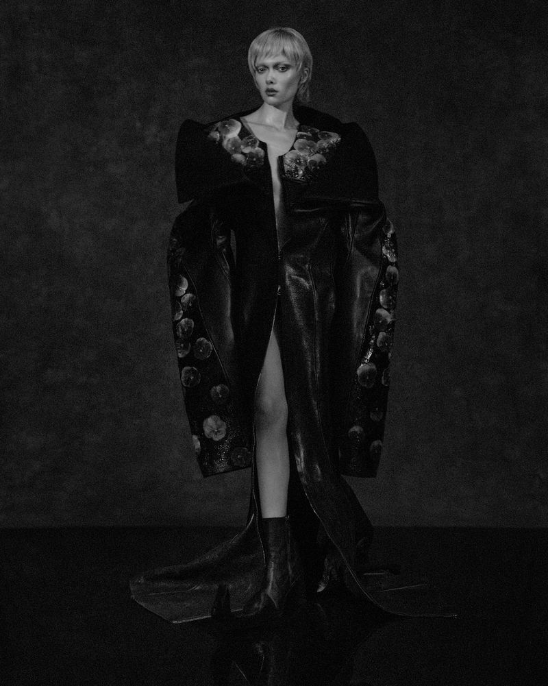 Monochrome fashion shot of a woman wearing an elaborate, long black coat with a floral pattern, standing against a dark backdrop.