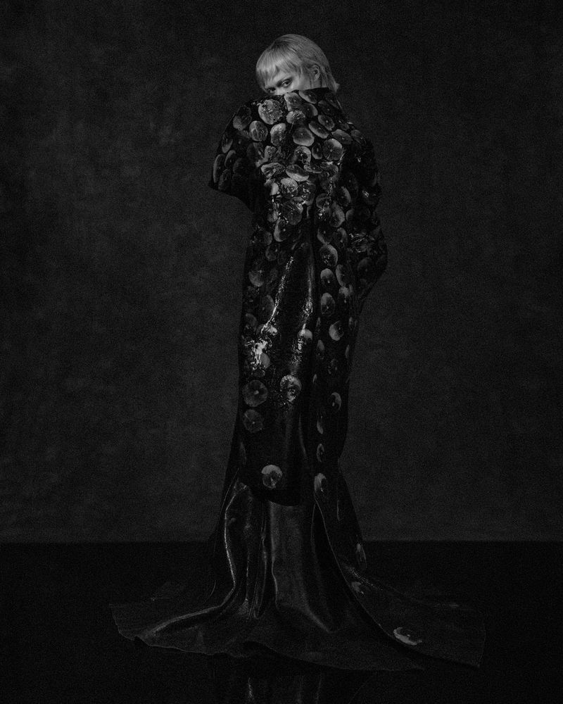 Monochrome portrait of a woman in a patterned robe, glancing back over her shoulder against a textured grey backdrop in an indoor setting.