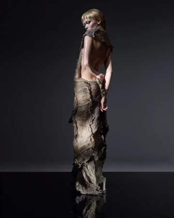Back view of slender adult female model wearing raw-edged earthy toned gown, with hands behind back. Shot on dark reflective surface.